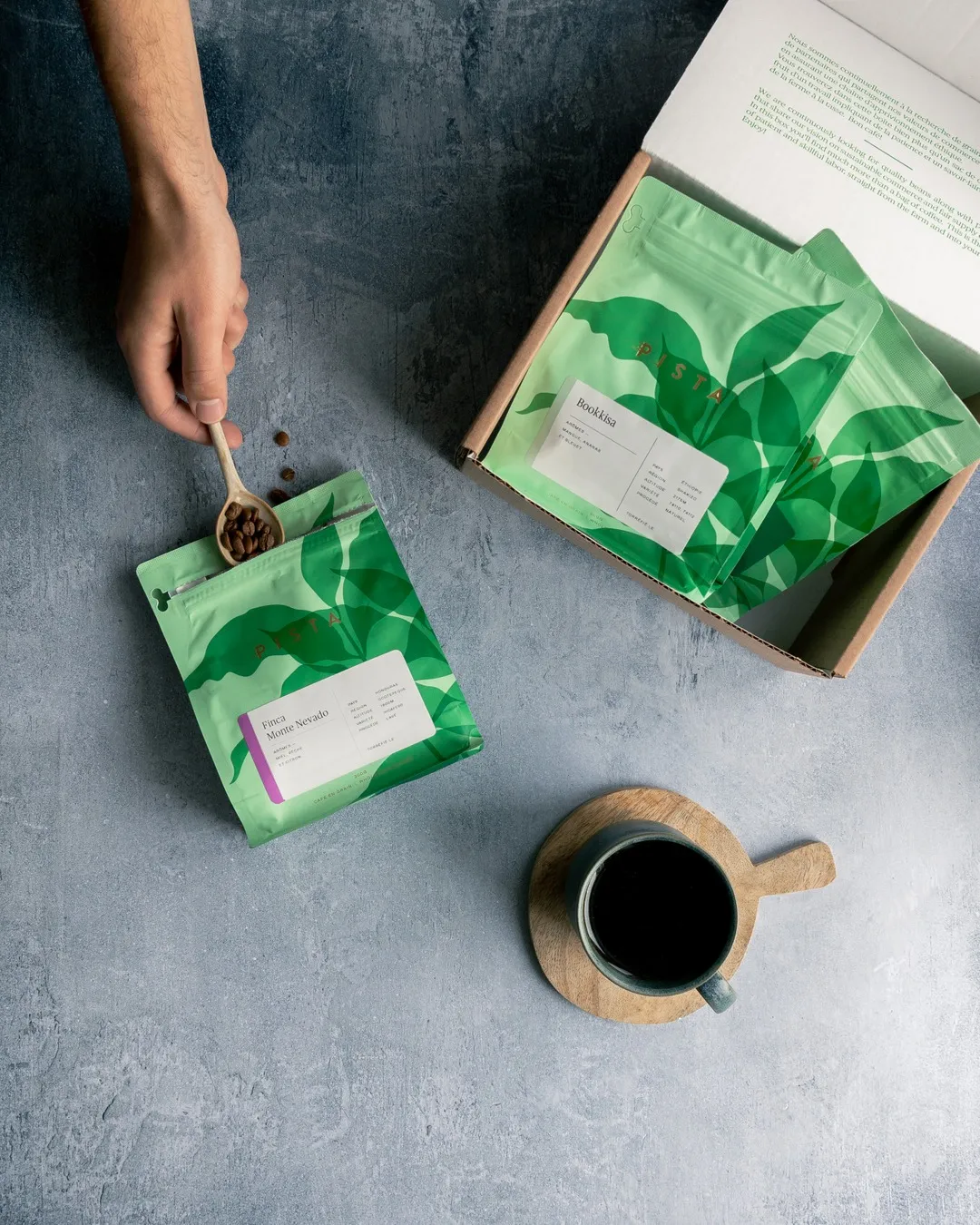 Une main verse des grains de café dans un sachet vert, près d'une boîte de sachets et d'une tasse de café.