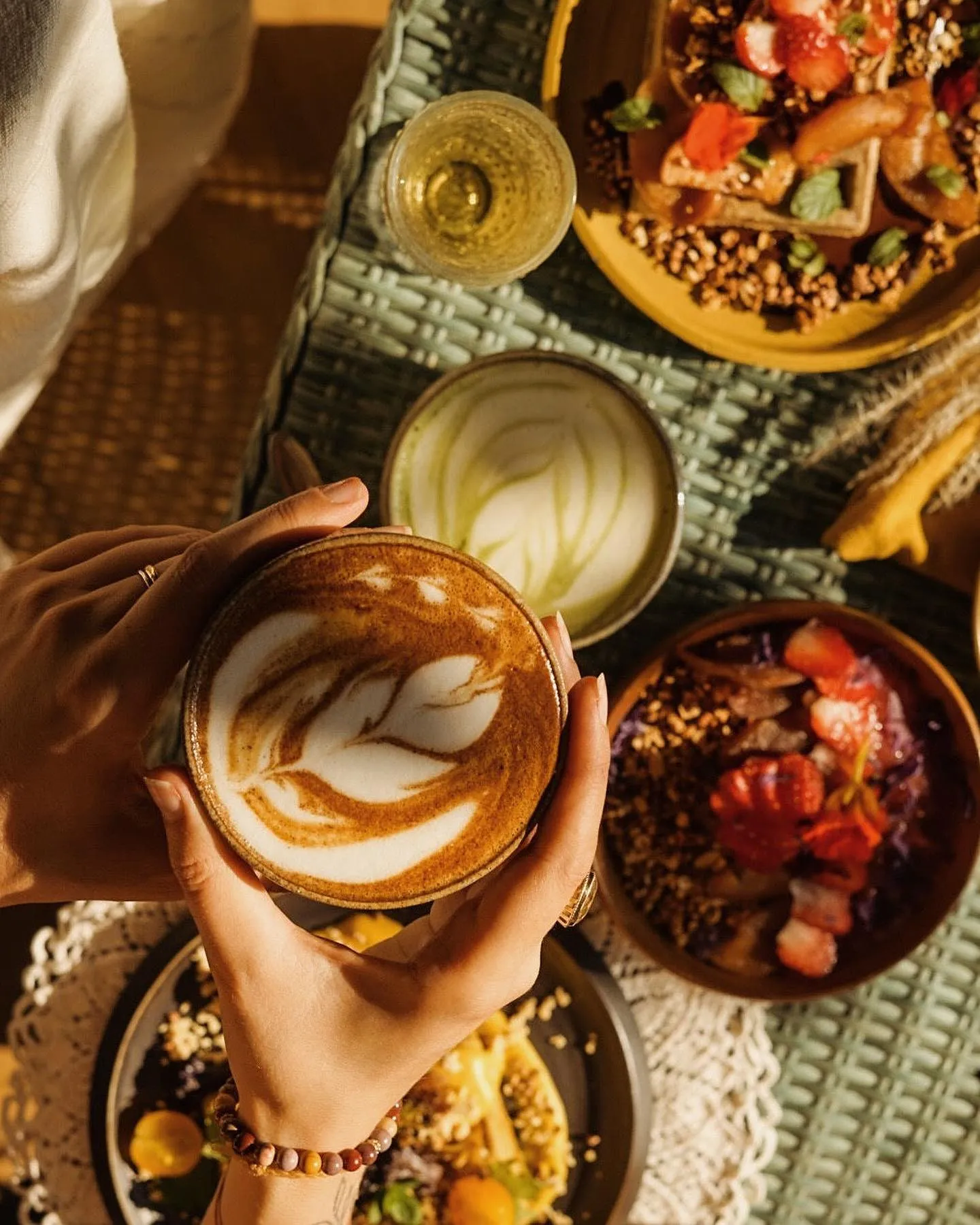 Des mains tiennent un cappuccino au lait dessiné, entouré de bols de fruits et de granola.