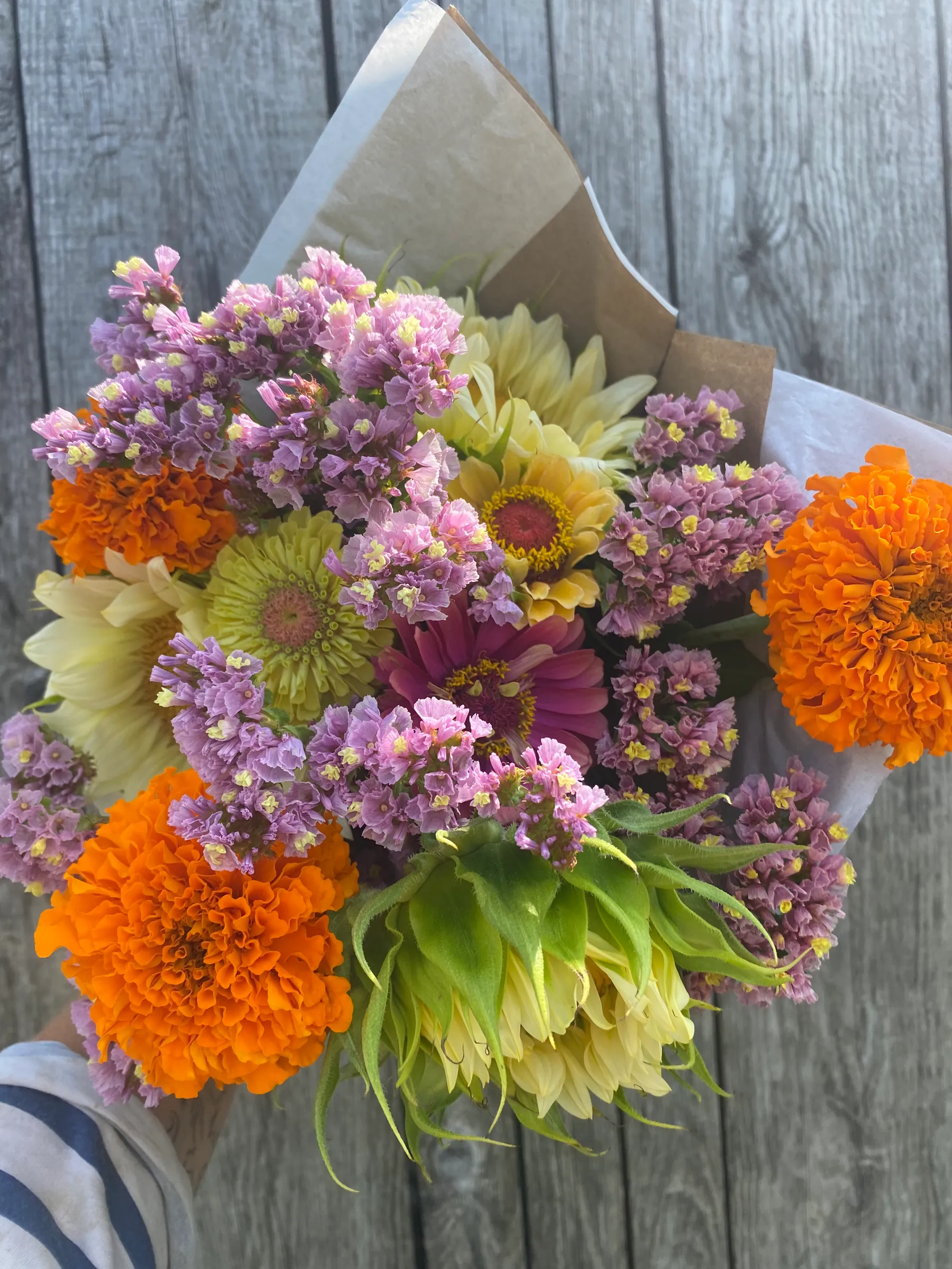 Bouquet de fleurs roses, jaunes et orange enveloppé dans du papier brun, posé sur des planches grises.